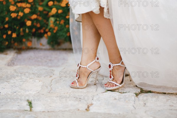 Bride's legs in stylish sandals with crystals with high heels peek out from under the wedding dress, close-up .