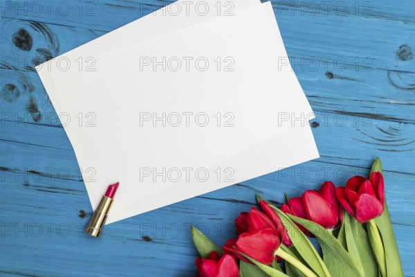 Red tulips, lipstick and a white sheet of paper for your greetings on the background of blue wooden board