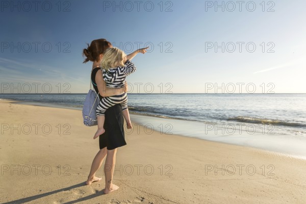 Young woman holding toddler daughter on her arm and pointing towards the ocean at the beach