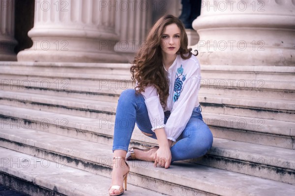 Young girl with brown hair and blue eyes, sits on old marble steps, dressed modern and looking at the camera