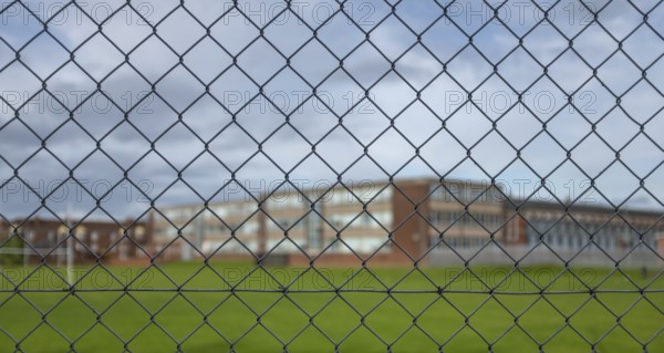 A Typical High School Building And Sports Field Under A Cloudy Sky With Focus On The Security Fence In The Foreground