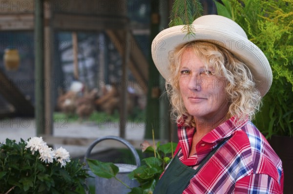 Side head-and-shoulders portrait of a middle-aged blonde woman with straw hat and gardener's apron in front of a chicken coop smiling friendly into the camera