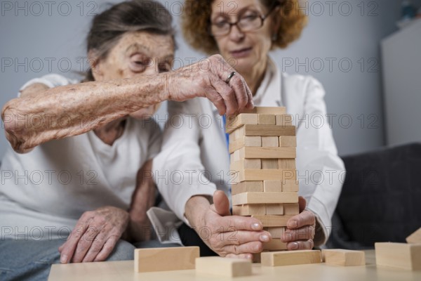 Senior woman practice skills, build wooden blocks, build tower and try not to let it fall, Jenga game. Old patient pull out block, place on top, support doctor during therapy dementia in house