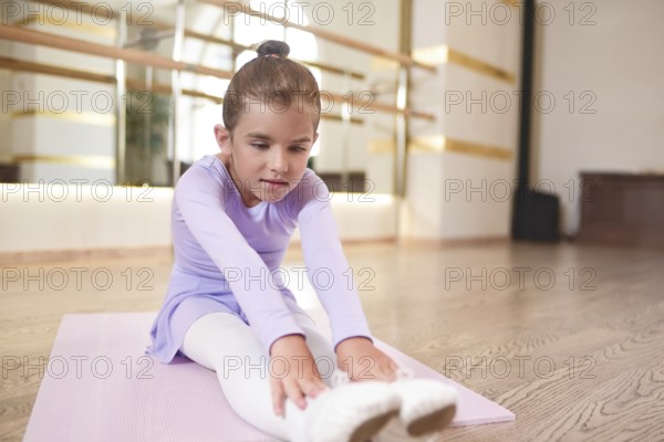 Adorable photo of little ballerina in a lilac dress and white tights stretching exercises in a hall with large mirrors