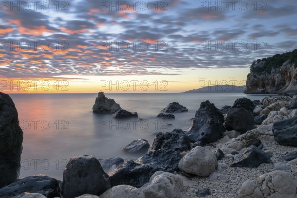A colorful sunrise at Cala Gonone beach with black and white rocks and boulders in the foreground