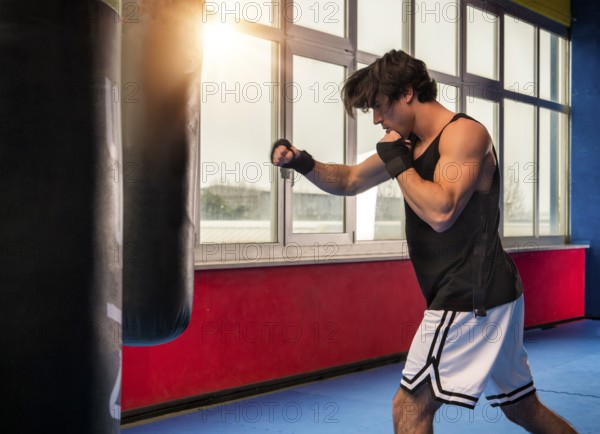 Side view of muscular sportive man in modern gym training with punching bag while boxing alone in back lit