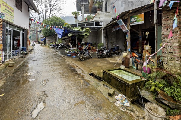 A narrow, wet road in a provincial area with parked motorbikes and colourful flags, road through a village in the Sa Pa area of Vietnam