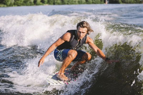 Beautiful long haired blond wakesurf in vest on board along waves of lake. Athletic male athlete wakes surfing in summer on river. Wakesurf water sport. Surfer on wave. Man surfing on the surfboard