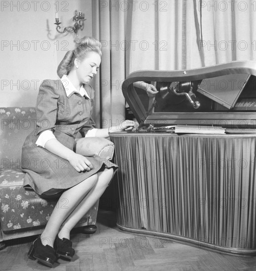 Woman at a gramophone in the 1940s. She has put a record on and is listening to the music. The records were made of fragile material and the speed of the record to sound as it should was 78 revolutions per minute. The gramophone is a luxury model with a satin fabric covering the sides. Sweden 1947. Kristoffersson ref AB22-2