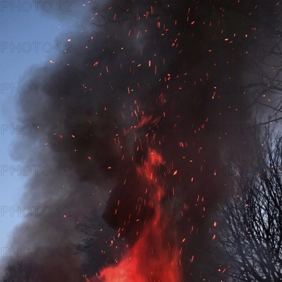 Crackling flames eat into the high pile of an Easter fire, dark smoke with flying sparks rises and darkens the surroundings. Square format. Crackling flames move through a high-piled heap of an Easter Saturday bonfire upwards, dark smoke with flying sparks rises up and obscures the environment. Square Format