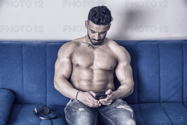 Handsome shirtless muscular bodybuilder man typing or surfing the internet with cell phone while laying on couch