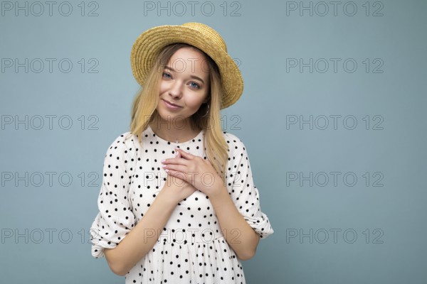 Foto von jung positiv glücklich schöne blonde Frau mit aufrichtigen Emotionen tragen Sommerkleid und Strohhut isoliert über blauen Hintergrund mit Kopierraum und honding Hände auf der Brust
