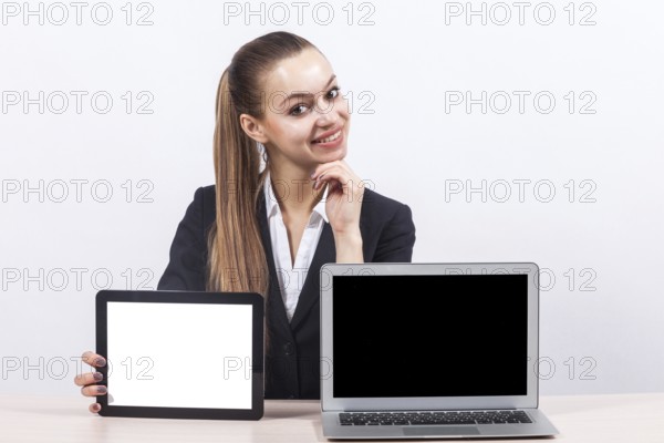 Attractive young caucasian businesswoman with ponytail in black suit sit at the office desk and show empty laptop and empty tablet, touching face, look at camera Isolated on grey