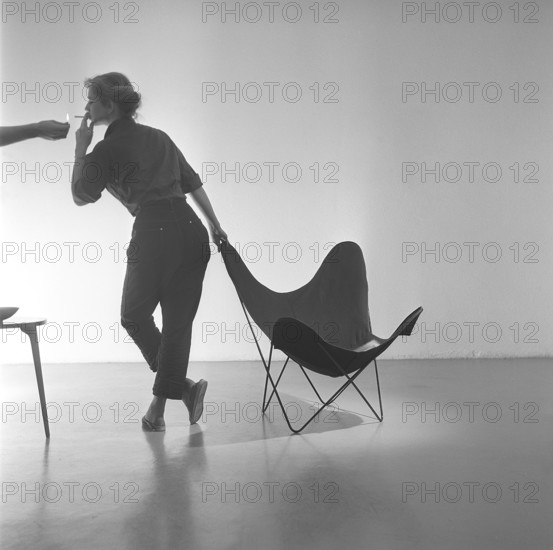 Home decor of the 1950s. A young woman is standing beside the popular Butterfly chair, also known as a BKF chair or Hardoy chair. The chair has its nickname due to the design. For many people the chair is a symbol of the youth rebellion against a generation of parents that wanted their children to sit up straight. The chair was created by two argentinian architects Juan Kurchan and Jorge Ferrari Hardoy, and spanish Antonio Bonet in the 1930s. It was named BKD but was considered hard to sell, probably because of its futuristic design. American Hans Knoll re introduced it 1947 and it became an international bestselling furniture. Sweden 1955. ref CV17-6