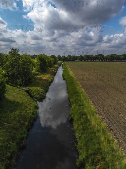 A quiet river runs between a planted field and a row of trees under a cloudy sky, aerial view of a river in the municipality of Hude (27798), Germany, Europe
