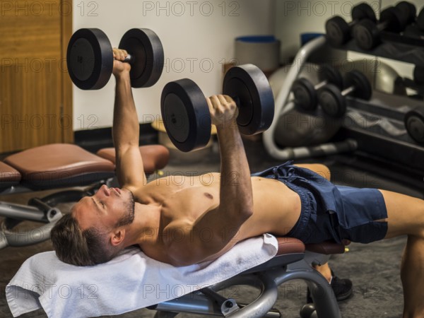 Muscular young man shirtless, lifting dumbbells training pecs on gym bench