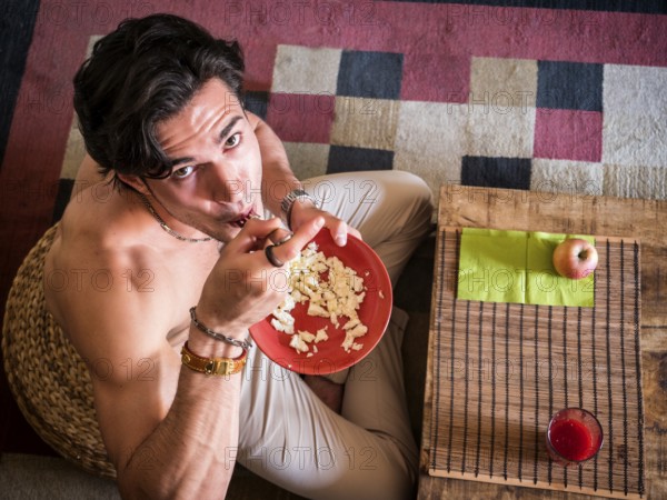 From above shot of happy muscular shirtless man sitting on floor having breakfast eating cereal, smiling at camera
