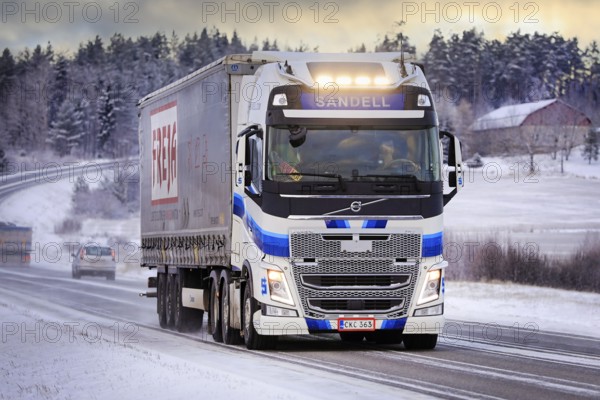 Blue white Volvo FH semi trailer truck Kuljetusliike Harri Sandell Oy with bright headlights on road 52 at winter dusk. Salo, Finland. Dec 27, 2021