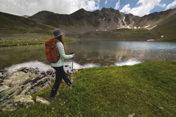 A woman with an orange backpack and a hat goes to a transparent lake in the mountains on a clear day