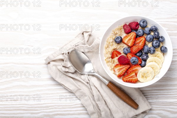 Oatmeal porridge with fruit and berries in bowl with spoon on white wooden background table top view, homemade healthy breakfast cereal with strawberry, banana, blueberry, raspberry. Space for text, food photography, food photography