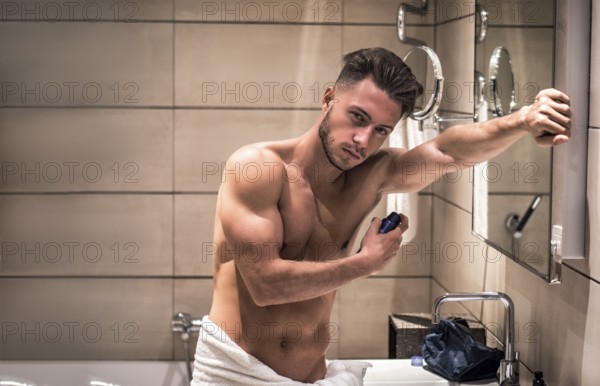 Handsome shirtless athletic young man in his home bathroom, spraying deodorant on armpits