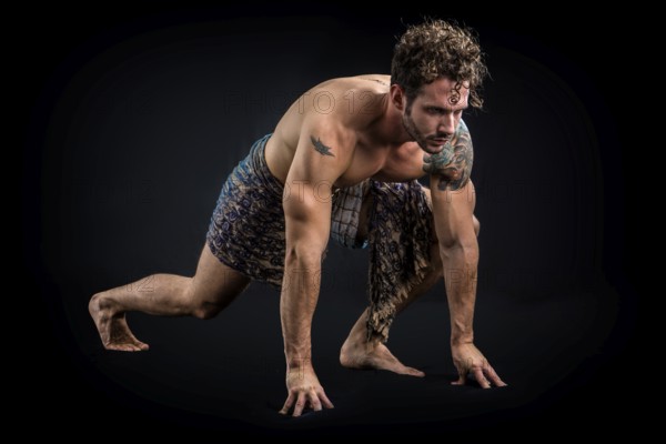 Handsome shirtless muscular man in briefs, covering with long elegant scarf, kneeling, isolated on black background in studio shot