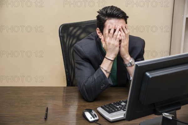 Desperate, worried young businessman sitting at his desk in front of his computer with his face in his hands and eyes closed