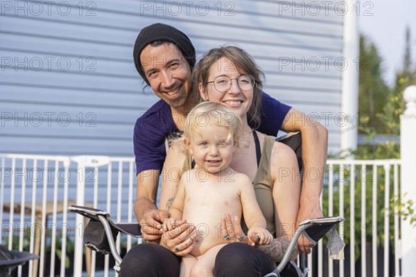 Happy young family pose for group portrait seen from the front. Mother sitting on chair with two year boy in lap and smiling father standing behind
