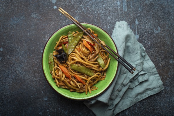 Asian dish stir fry udon noodles with chicken, vegetables and mushrooms in green ceramic bowl with wooden chopsticks on rustic dark blue concrete background from above, Chinese or Thai fast food, food photography