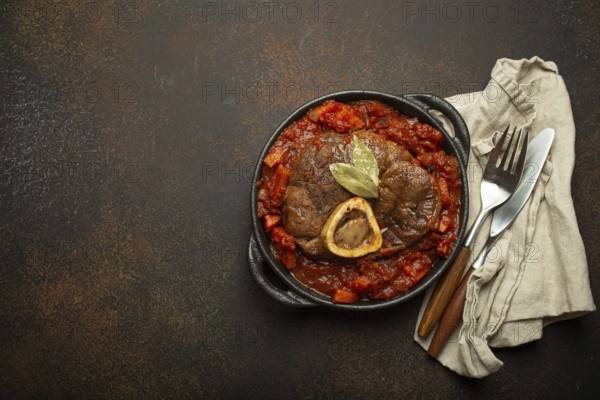 Traditional Italian dish Ossobuco all Milanese made with cut veal shank meat with vegetable tomato sauce served in black casserole pan top view on rustic brown background, copy space, food photography, food photography