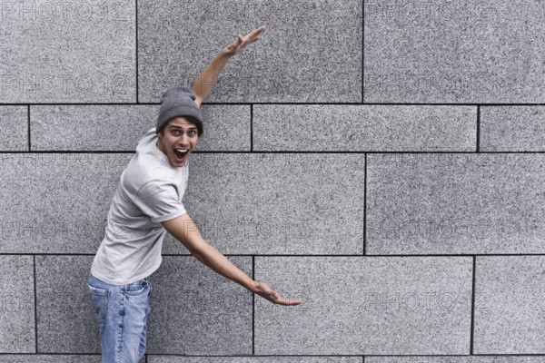 Cheerful guy dressed in gray t-shirt, sunglasses and hat at the street, show a big sale, or nice offer. Advertise concept. People, travel and tourism, man on city street show with his hands something over gray wall background