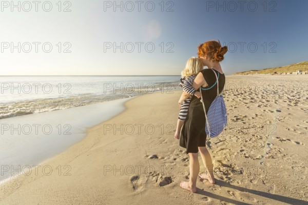 Woman is standing on a lonely beach with her child in her arms, looking into the sunset