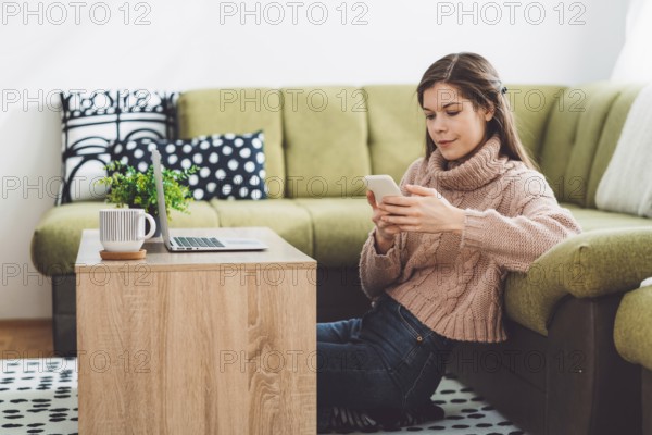 Young caucasian woman with long brown hair in pastel pink sweater and jeans working from home on her laptop and phone. Cheerful woman student studying at home