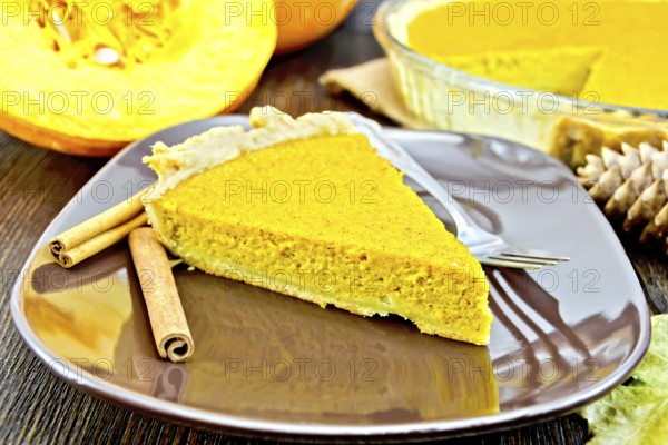 A piece of pumpkin pie in a brown plate with a fork and cinnamon, pumpkin, cones and maple leaf on the background of dark wood planks