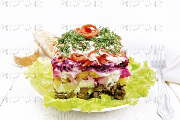 Puff salad with beef, boiled potatoes and beets, pears, spicy carrots, seasoned with mayonnaise and garnished with dill on a green lettuce in a plate, towel, bread on light wooden board background
