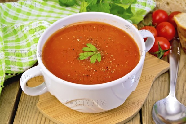 Tomato soup in a white bowl, green napkin, spoon, tomato, parsley, bread on a wooden boards background
