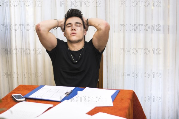 Tired, exhausted or despondent young man sitting at a table with a folder of paperwork leaning forwards with eyes closed