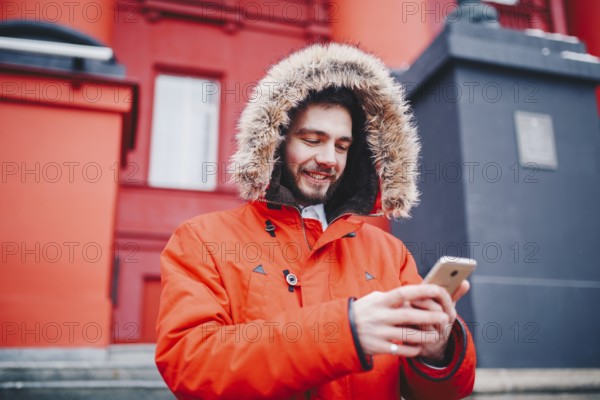 Handsome young male student with toothy smile and beard stands on red wall background, facade of educational institution in red winter jacket with hood with fur, Uses finger on screen of mobile phone