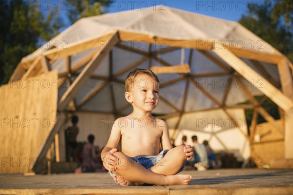 Theme is yoga and children. Caucasian Boy child sitting barefoot cross-legged in lotus position on wooden floor the background of gathering place of people. In summer sunset. Dressed in denim shorts