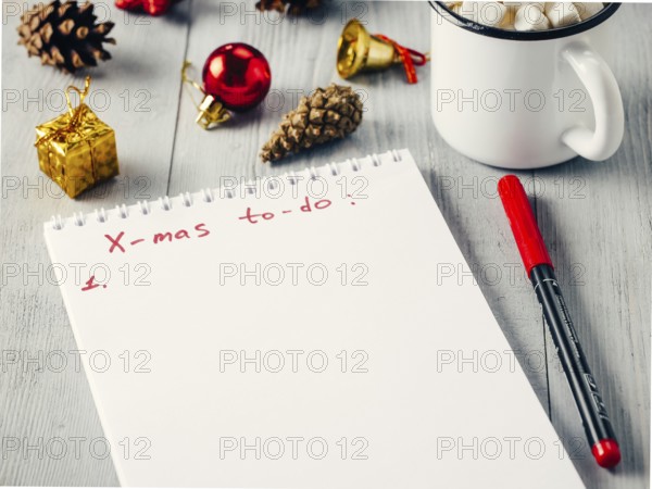 Christmas gifts shopping planning. Make shopping or to-do list for Christmas. Notebook, mug hot chocolate with marshmallows, New Year's decoration and pine cone on gray wooden background