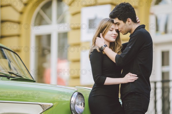 Young multiracial couple, male and female lovers heterosexual people students. Beautiful models posing standing near a retro car in the city. Dressed in black clothes