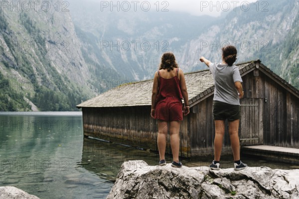 Two girls standing on top of a rock and wooden cottage in beautiful lake in the Alps with misty mood
