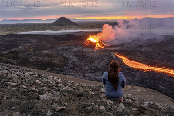 A woman sits on a mountain and looks at a glowing volcano, Grindavik, Sudurnes, Iceland, Europe