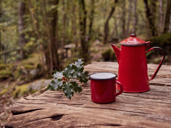 Christmas breakfast in the woods. Sprig of holly next to red mug and vintage coffee pot on wooden picnic table in outdoors