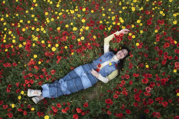 Young happy girl resting in the field of red poppy flowers. Drone aerial of woman in spring