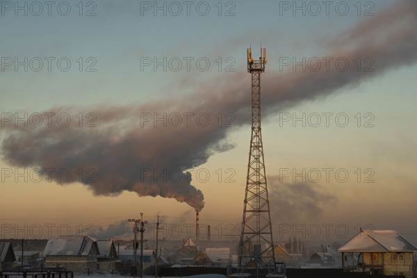 Smoke rises from the chimney of the boiler room over the city and the cell tower is in the foreground. Russia, Novosibirsk region, air temperature -41 degrees Celsius