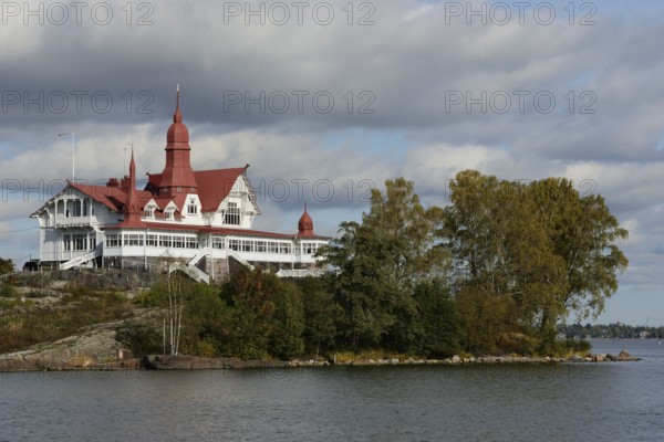 This image captures a historic seaside building with distinctive red roofs, nestled on a small island by the waterfront. The architecture features a blend of Victorian and Nordic design elements, highlighting the cultural heritage of the region. Surrounded by autumn foliage and calm waters, the building stands out as an iconic landmark, offering a glimpse into Finland's rich architectural history. The setting, with its serene coastal landscape and overcast sky, adds to the picturesque and tranquil atmosphere of the scene