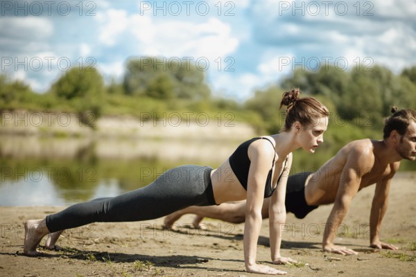 Woman and man in plank positions on a sandy area by a lake, emphasizing core strength