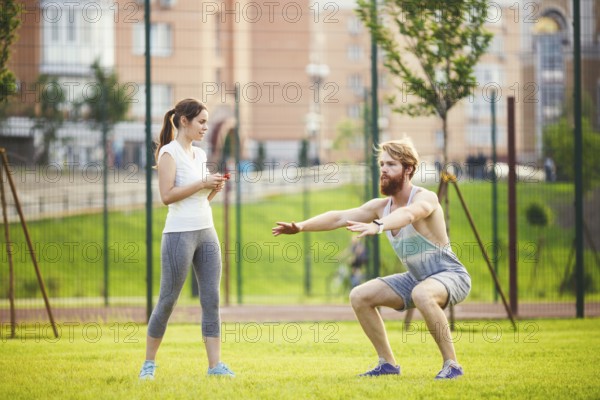 Team work in sports. female trainer training man with beard in park on green grass, lawn at sunset. The girl holds phone in hand and uses stopwatch, guy does exercises for the buttocks of sitting down