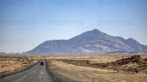 A car drives on a dusty road towards a towering mountain in the background, By car through the Sahara in Algeria